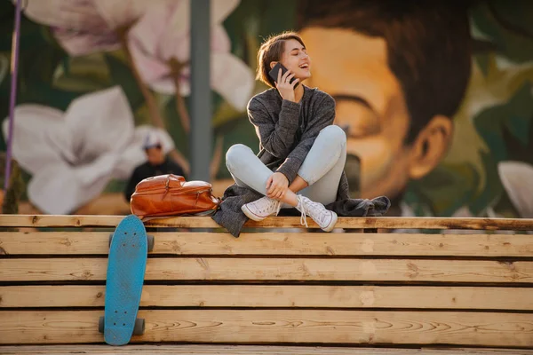Jovem com um skate está descansando em um banco de 2 estágios, fazendo uma chamada . — Fotografia de Stock