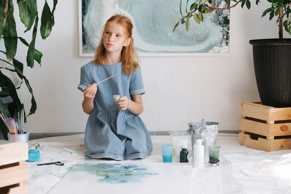 Adolescente ruiva menina está pintando com escova em tela grande no chão em uma oficina . — Fotografia de Stock