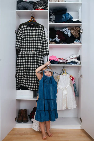 Kleines lustiges Mädchen probiert neues Kleid vor offenem Kleiderschrank voller Kleider an — Stockfoto