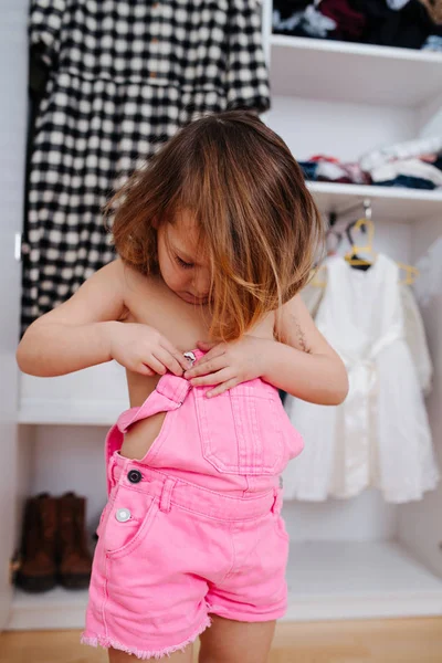Little girl trying on new overalls in front of open wardrobe full of clothes — Stock Photo, Image