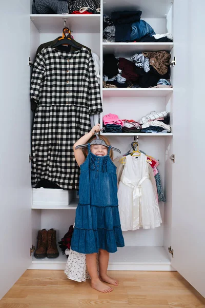 Little jolly girl trying on new dress in front of open wardrobe full of clothes