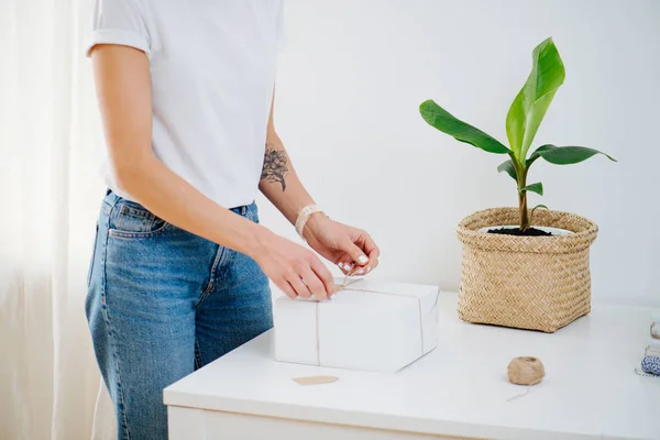 Woman in casual clothes is tying postal cardboard box, attaching tag at home — Stock Photo, Image