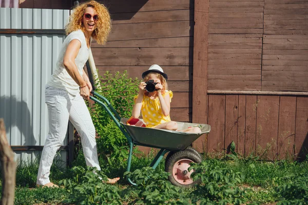 Mother driving a wheelbarrow, while her daughter with camera riding in it