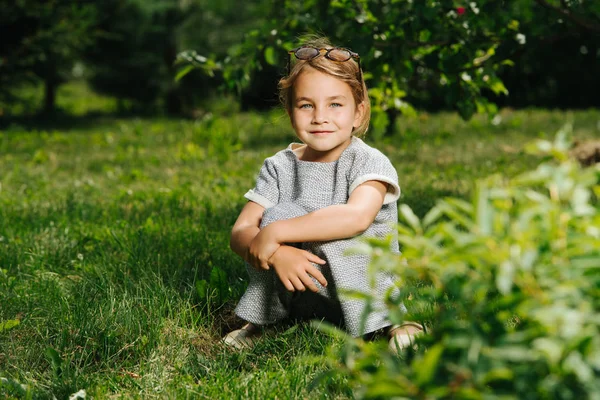 Lycklig liten flicka som sitter på ett gräs i trädgården. Njuter av sommardagen — Stockfoto
