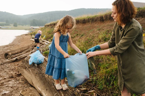 Madre con sus hijas recogiendo botellas en bolsas de basura en una orilla del río — Foto de Stock