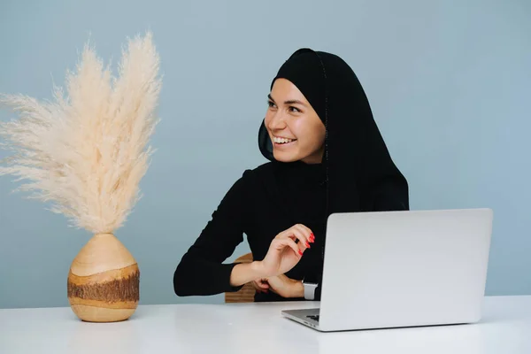 young smiling muslim woman look away and working on a laptop, sitting at a table