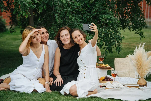 Four young women are taking selfie during picnic on a lawn under tree