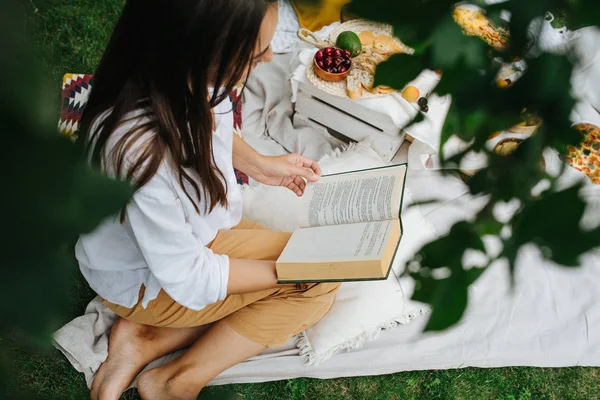 Jonge vrouw is het lezen van een boek, zittend op haar benen, op een witte doek bij picknick — Stockfoto