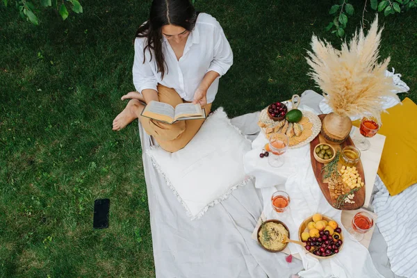 Mujer joven está leyendo un libro, sentada sobre sus piernas, en un paño blanco en el picnic —  Fotos de Stock