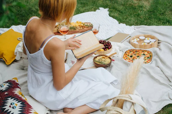 Genç kadın piknikte beyaz bir bezin üzerinde, bacaklarının üzerinde oturarak kitap okuyor. — Stok fotoğraf