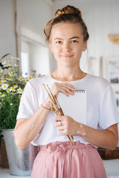 Portret cute uśmiechnięta młoda kobieta trzyma notebooka i szczotki w dłoniach — Zdjęcie stockowe