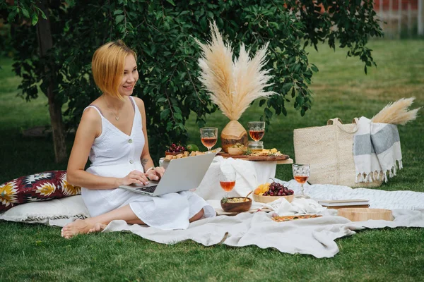 Dizüstü bilgisayar ile genç kadın, piknikte beyaz bir bez üzerinde, bacaklarının üzerinde oturan — Stok fotoğraf