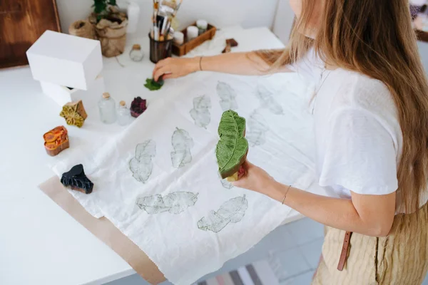 나무 스탬프가 있는 직물에 녹색 잎 모양의 프린트를 만드는 젊은 여성. — 스톡 사진