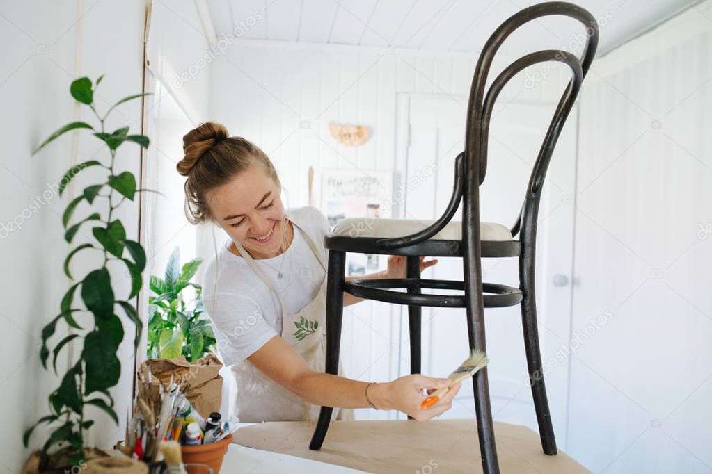 young woman varnishing, renewing an old wooden chair in a bright room at home