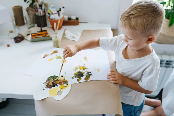 Cute first grade child boy is painting on paper sheet with brush at home.