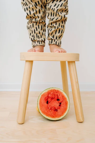 Little boy\'s feet standing on stool. Red watermelon half slice is under it.