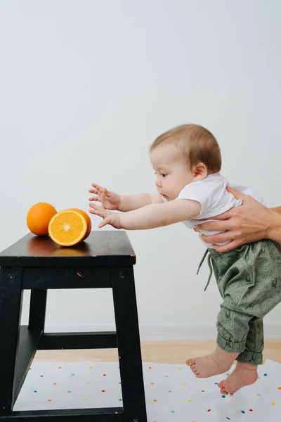 Cute baby in his mother's hands trying to reach oranges — Stock Photo, Image