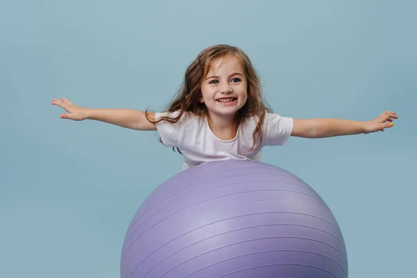 小女孩试图爬上瑜伽球，没有手在蓝色背景 — 图库照片