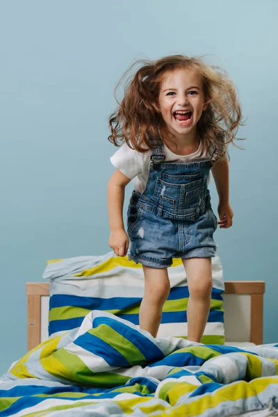 Little girl is playing actively, jumping in bed over blue background. — Stock Photo, Image