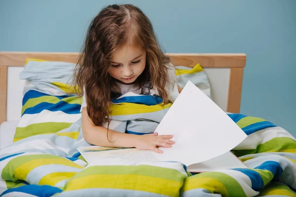 Klein meisje is het lezen van boek in bed voordat u gaat slapen. Over blauwe achtergrond. — Stockfoto