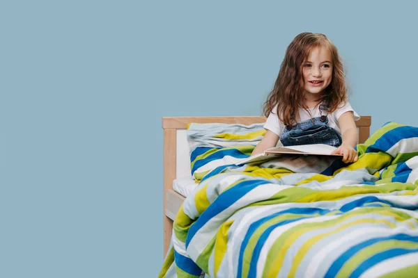 Little girl is reading book in bed before going to sleep. Over blue background. — Stock Photo, Image