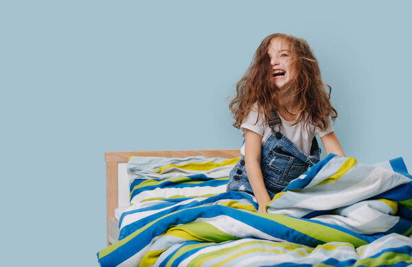 Little girl is playing actively, frolicing in bed over blue background.