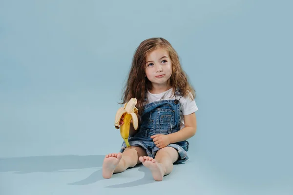 Menina da criança comendo banana, retrato de estúdio no fundo azul — Fotografia de Stock