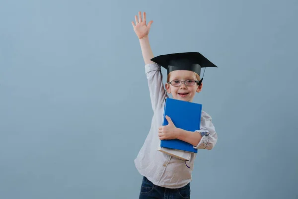 Der rothaarige Erstklässler mit Brille und Studentenmütze hebt den Arm über Blau — Stockfoto