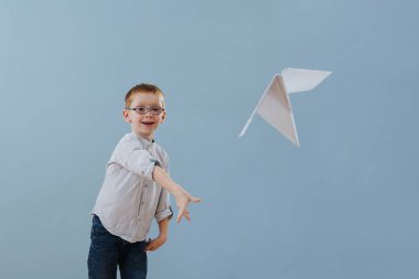 Sevimli küçük kızıl çocuk mavi arka planda uçmak için kağıt bir uçak fırlattı.
