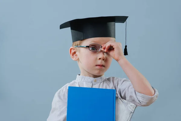 Рыжий первоклассник в студенческой шляпе с книгами в руках чинит очки. — стоковое фото