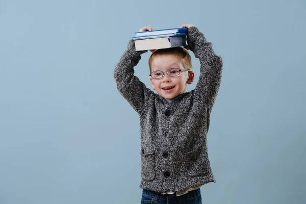 Carino poco zenzero ragazzo in occhiali tenendo mucchio di libri su la sua testa oltre blu — Foto Stock