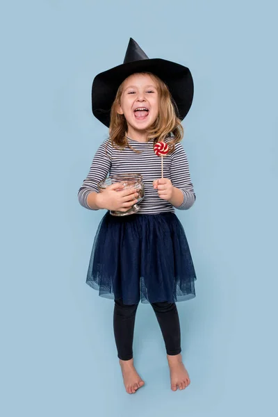 Portrét malé holčičky v čarodějnickém klobouku držící lízátko a sklenici plnou lahůdek — Stock fotografie