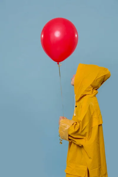 Mały chłopiec w żółtym płaszczu przeciwdeszczowym z czerwonym balonem na niebieskim tle — Zdjęcie stockowe