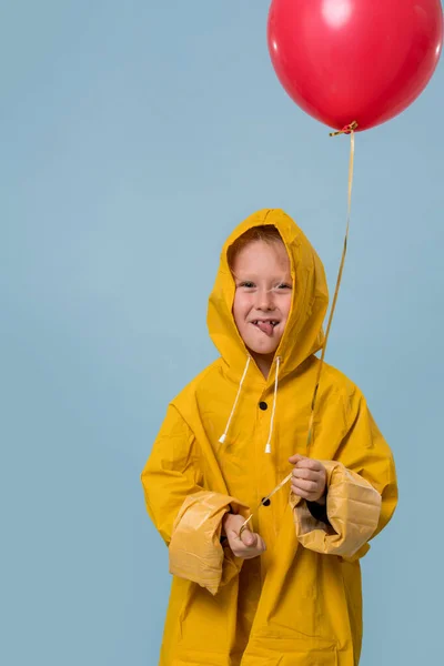 Little boy in yellow raincoat with red balloon over blue background
