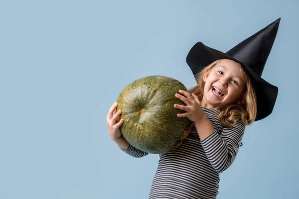 Portrét malé holčičky v čarodějnickém klobouku a černém oblečení s dýní přes modrou — Stock fotografie