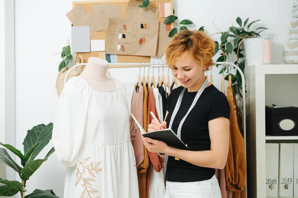 A young dressmaker in a workshop writes in notepad, she is working on a new dress design