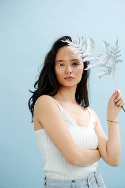 Yüzünün yanında eğreltiotu tutan güzel bir esmerin portresi. Beyaz vitiligo kirpikleri var. Vücudu yarı yarıya değişmiş. Mavi arkaplan.