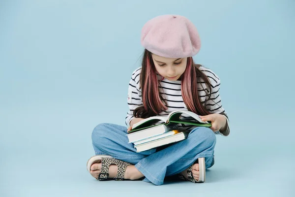 Fashionable little girl in a pink french beret sitting cross-legged on the floor, reading book over blue backgorund.
