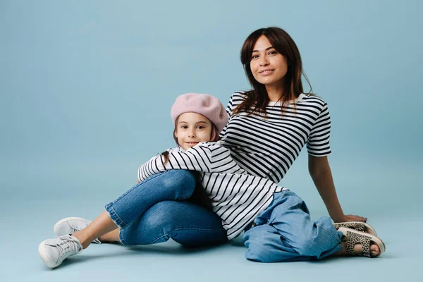 Two fashion lovers, mom and daughter wearing same clothes. They have jeans and black and white striped shirt on them. Mom sitting on the floor and leaning on the floor. Dau leaning on her.