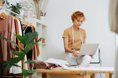 Stüdyoda bir masanın üzerinde oturan ve elinde dizüstü bilgisayarla çalışan bir tasarımcı.