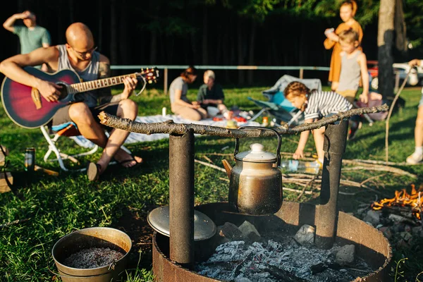 Friends with families having picnic by the campfire on a beautiful day. Iron teapot hanging over live coals. Man playing guitar nearby.