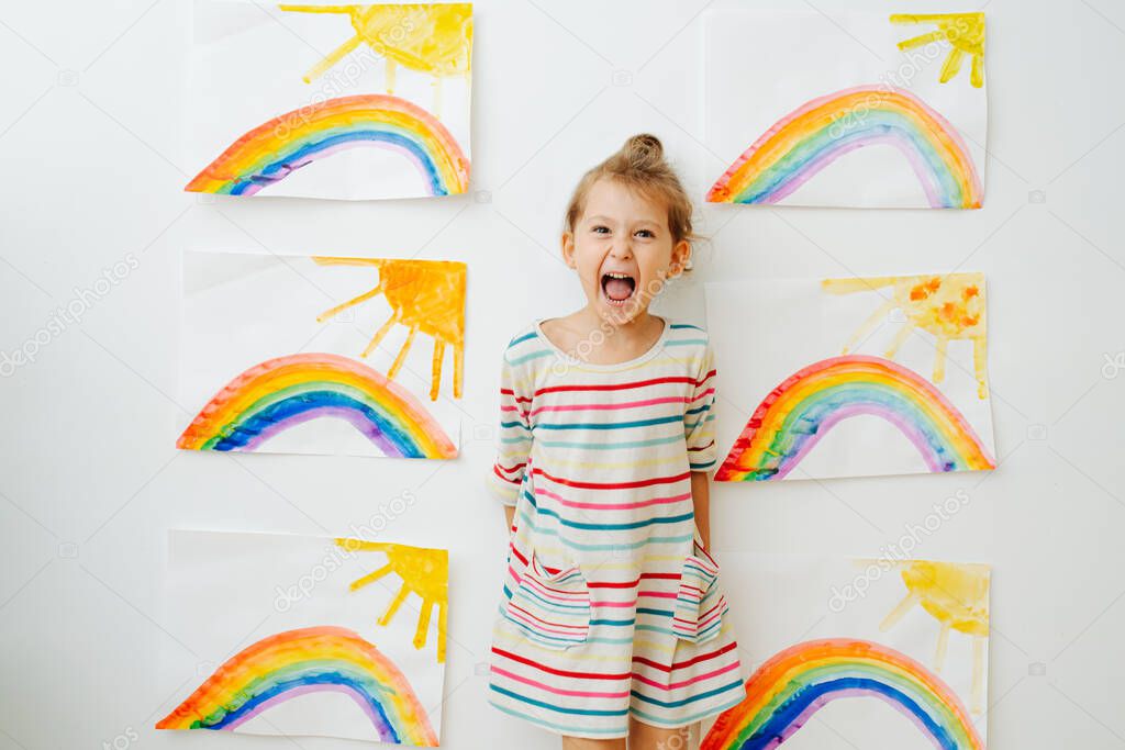 Hyperemotional child cheering to the amount of rainbow and sun paintings on the wall. Girl wears white dress with colorful stripes.