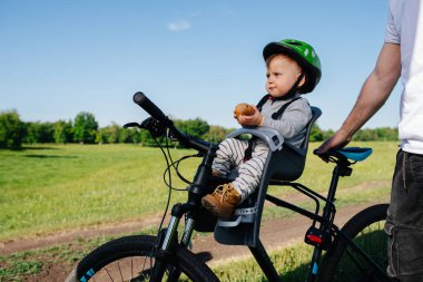 Huysuz suratlı bebek bisiklette emniyet sandalyesinde oturuyor, elinde armut tutuyor. Babası yakınlarda duruyor, aracı destekliyor..