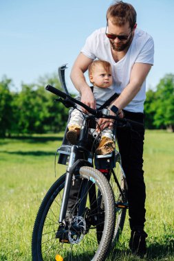Özenli bir baba, çocuğunu dikkatlice ön bisiklet bebek koltuğuna bağlıyor. Omuzlarına emniyet kemeri takıyordu. Yeşil kırsal alanda. Çocuk işleme pek düşkün değil..