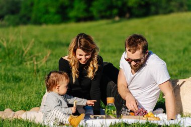 Ebeveynler ve küçük oğulları kırsalda piknik yapıyorlar. Yeşil bir tarlada battaniyenin üzerinde oturuyorlar..