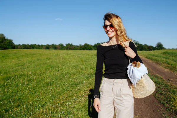 在绿地的乡间 一个戴着太阳镜的金发女人站在一条小径上 满脸笑容 她拿着装有野餐食物的袋子 — 图库照片