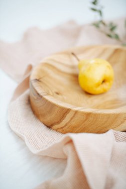 İçinde taze, sarı bir armut olan el yapımı ahşap bir kâse. Tabak pürüzsüz ve verniklenmiş, beyaz bir yüzeyde bir peçetenin üzerinde duruyor. Kurumuş dal bu kompozisyonu tamamlar. Seçici odak.