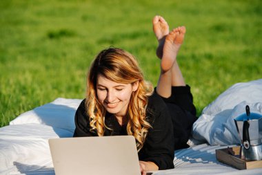 Güzel bir kadın laptopta çalışıyor, açık havada yatakta uzanıyor, güneşin tadını çıkarıyor.