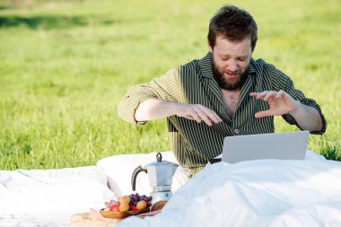 Adam laptopuyla çalışırken hapşırıyor, çimenlikteki yatakta oturuyor.