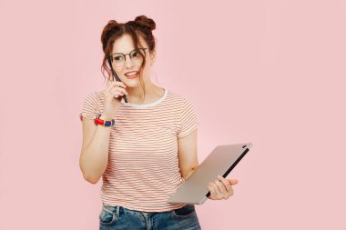 Genç meşgul bir kadın telefonla konuşuyor ve dizüstü bilgisayarını pembe arka plan üzerinde tutuyor. Çizgili gömlek ve kot pantolon giyiyor..